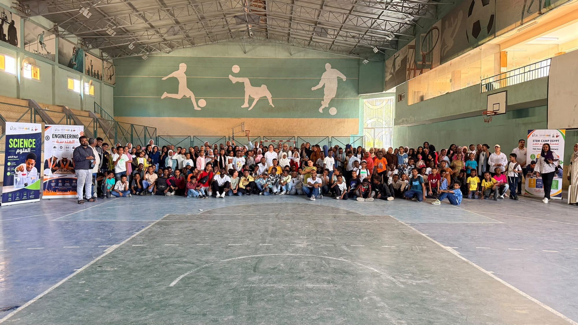 Children and facilitators gathered together in a sports hall at the end of a STEM camp day, between roll-up banners for the science and engineering stations.